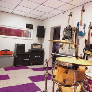 Foto von Aufnahmeraum 1 im StudioCarbon Tonstudio - Wiener Neudorf bei Mödling. Zu sehen sind ein Schlagzeug, mehrere Gitarrenverstärker sowie einige Gitarren an Wandhalterungen. Sicht auf das Regiefenster